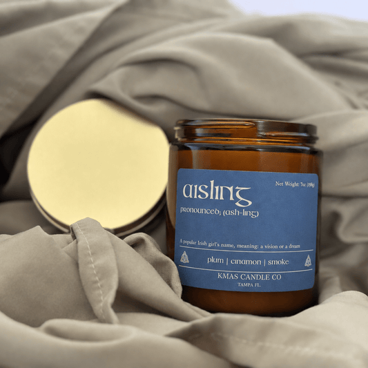 Aisling | Wooden Wick Candle - KMAS Candle Co-Candles-KMAS Candle Co-An amber glass candle jar with a blue label reading 'aishin' and 'KMAS CANDLE CO' sits on draped taupe fabric, with a gold-toned metal lid placed beside it.