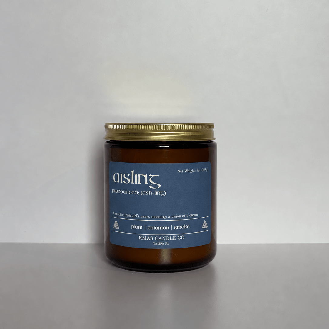 Aisling | Wooden Wick Candle - KMAS Candle Co-Candles-KMAS Candle Co-An amber glass jar candle with a gold metal lid and a blue label reading 'aisling' and 'plum | cinnamon | smoke', photographed against a plain light gray background.