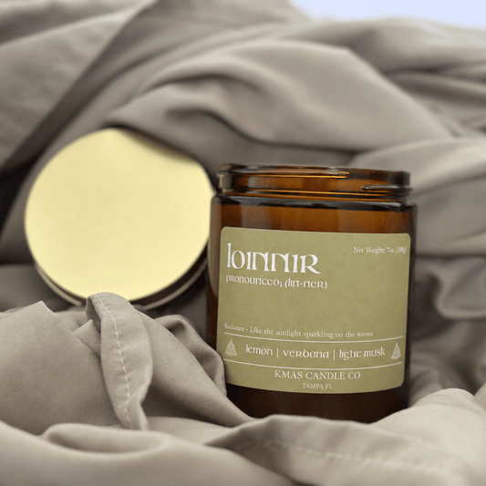 Loinnir | Wooden Wick Candle - KMAS Candle Co-Candles-KMAS Candle Co-An amber glass candle jar with a beige label reading 'lunnir' and 'Klagenfjord', placed on draped taupe fabric with its gold-toned lid beside it.