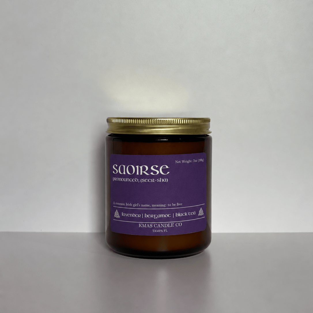 Saoirse | Wooden Wick Candle - KMAS Candle Co-Candles-KMAS Candle Co-A single amber glass jar candle with a gold metal lid and a purple label reading 'saoirse', photographed against a plain light gray background.