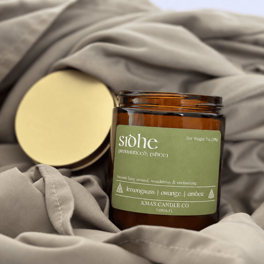 Sidhe | Wooden Wick Candle - KMAS Candle Co-Candles-KMAS Candle Co-An amber glass candle jar with a muted green label reading 'stöhe' and 'KMA Candle Co' sits on draped taupe fabric, with its gold lid placed beside it.