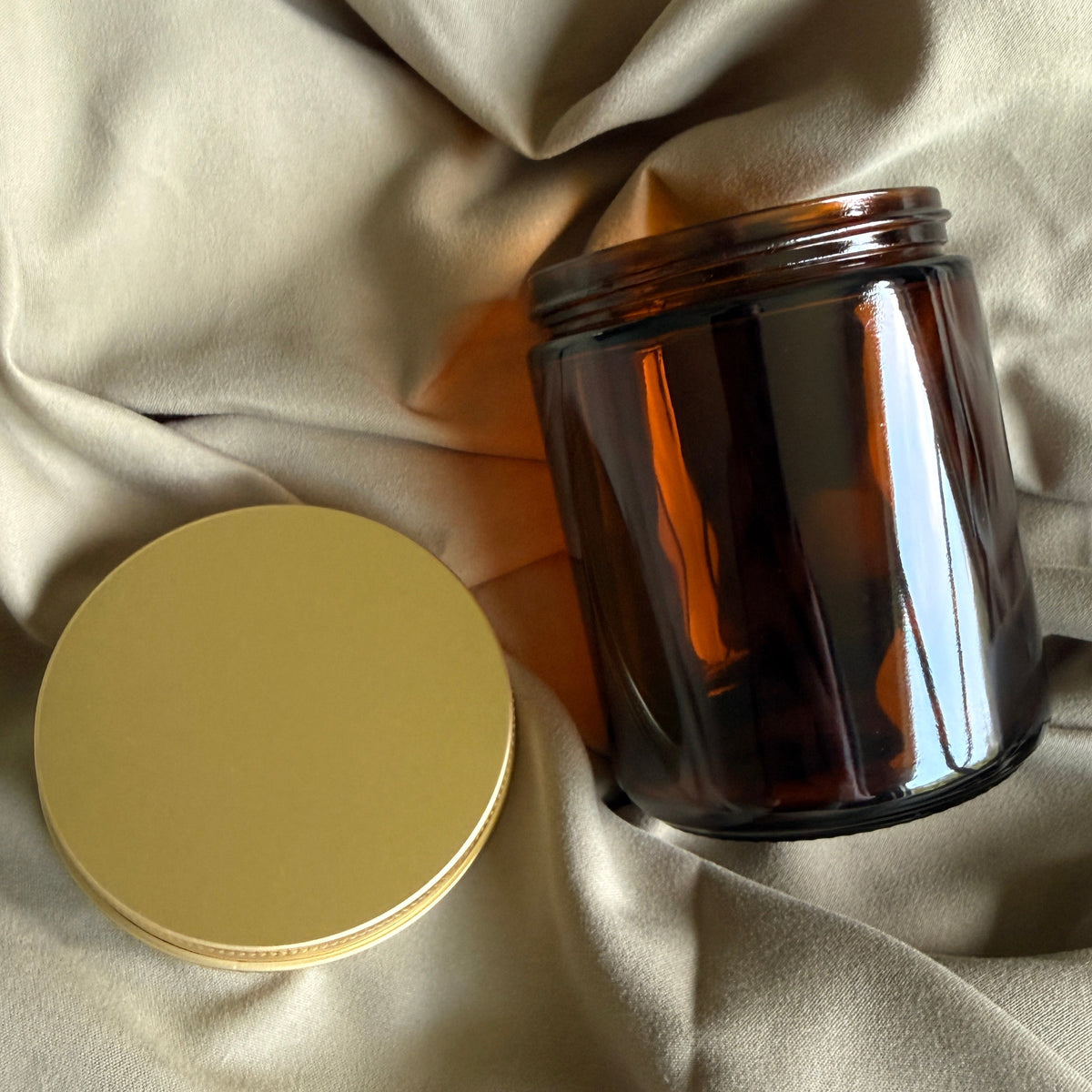 An amber glass jar candle with its gold metal lid resting beside it on a draped beige fabric background.
