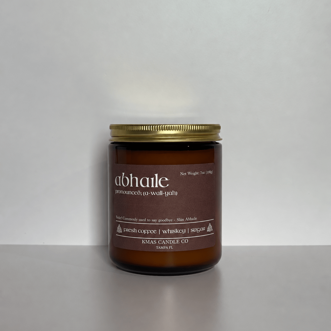 Abhaile | Wooden Wick Candle - KMAS Candle Co-Candles-KMAS Candle Co-An amber glass jar candle with a gold metal lid and a dark brown label reading 'abhaile' and 'pomegranate & wolf goby', photographed against a plain light gray background.
