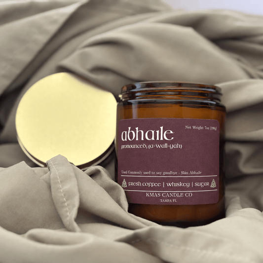 Abhaile | Wooden Wick Candle - KMAS Candle Co-Candles-KMAS Candle Co-An amber glass candle jar with a mauve label reading 'abhaile' and 'RMS Candle Co' sits on draped taupe fabric next to a gold metal lid.