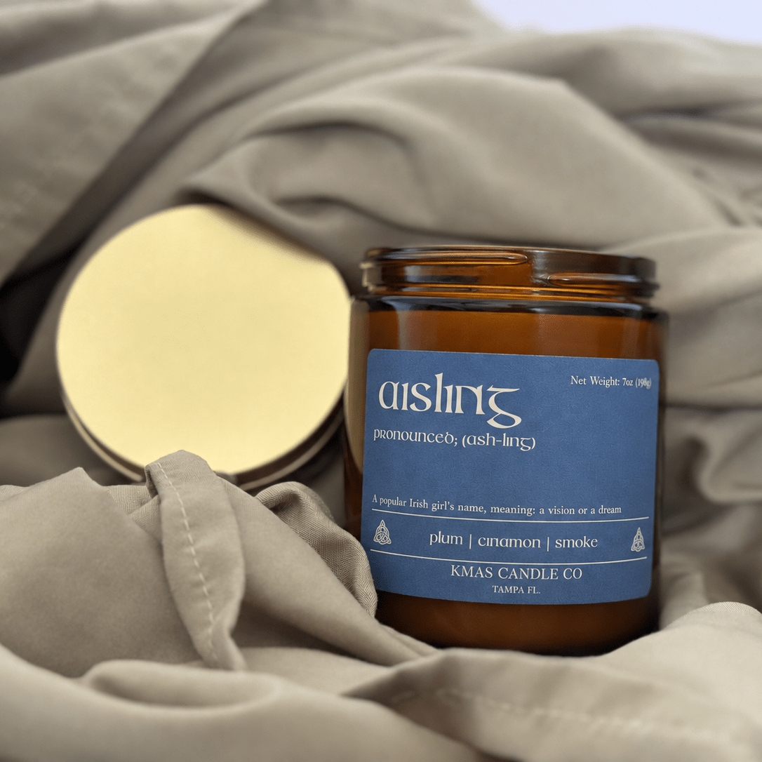 Aisling | Wooden Wick Candle - KMAS Candle Co-Candles-KMAS Candle Co-An amber glass candle jar with a blue label reading 'aishin' and 'KMAS CANDLE CO' sits on draped taupe fabric, with a gold-toned metal lid placed beside it.