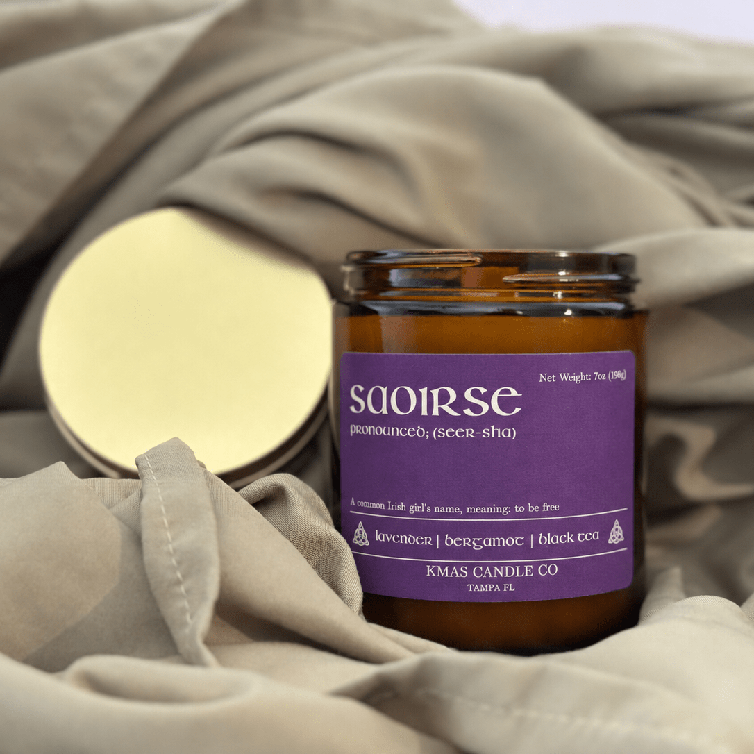 Saoirse | Wooden Wick Candle - KMAS Candle Co-Candles-KMAS Candle Co-An amber glass jar candle with a purple label reading 'soirse' and 'KMAS CANDLE CO', placed on draped beige fabric with its gold-toned lid beside it.