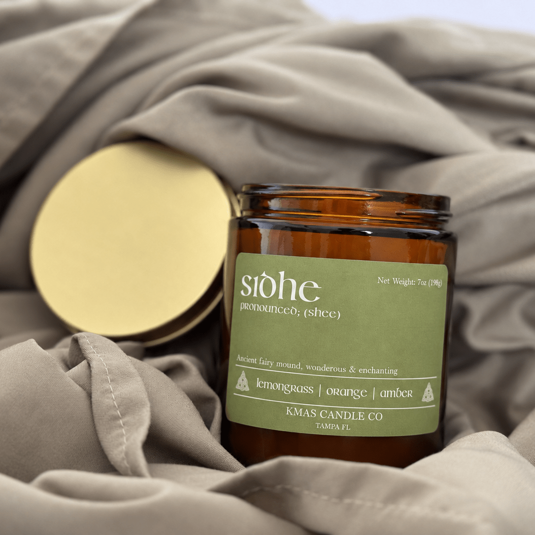 Sidhe | Wooden Wick Candle - KMAS Candle Co-Candles-KMAS Candle Co-An amber glass candle jar with a muted green label reading 'stöhe' and 'KMA Candle Co' sits on draped taupe fabric, with its gold lid placed beside it.